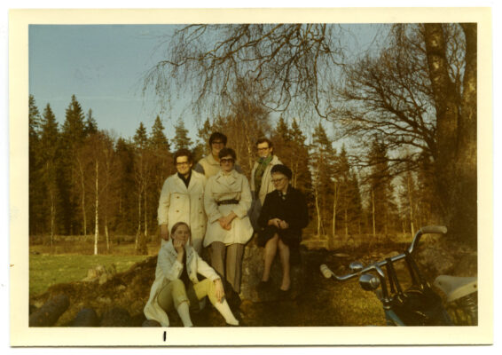 6 kvinnor sitter och står på en liten kulle. I bakgrunden skymtar ett fält och skog. I förgrunden skymtar styret på en cykel.