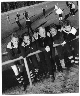 Fem fotbollsspelare lutar sig över ett staket för att låta sig fotograferas. I bakgrunden skymtar pågående fotbollsträning.
