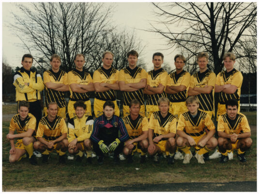 17 fotbollsspelare i svargula matchställ uppställda på gräsmatta. I bakgrunden skymtar några träd utan löv.