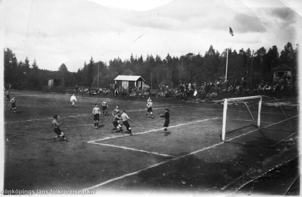 Foto taget snett ovan kortlinjen över fotbollsmatch. Längre bort syns spridd publik på bänkar och bergshällar.