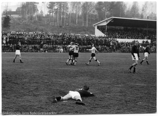 Fotbollsmatch. I förgrunden ligger en spelare i gräset. I bakgrunden syns en fullsatt läktare.