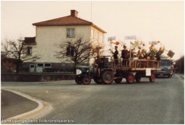 En grupp människor med ballonger och björkris körs på ett flak bakom en liten traktor.