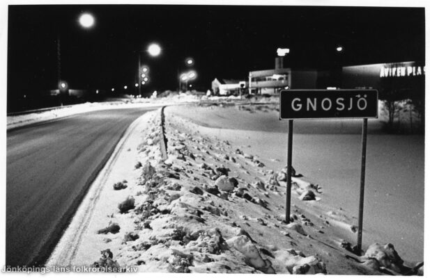 Vinterkvällsbild över väg. Vid sidan av vägen en skylt med texten "GNOSJÖ". I bakgrunden bebyggelse.