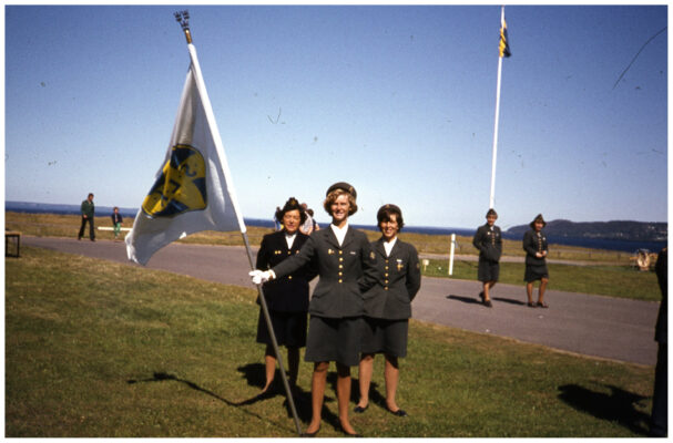 Tre kvinnor i Lottauniform uppställda för foto. Den främre håller en fana.