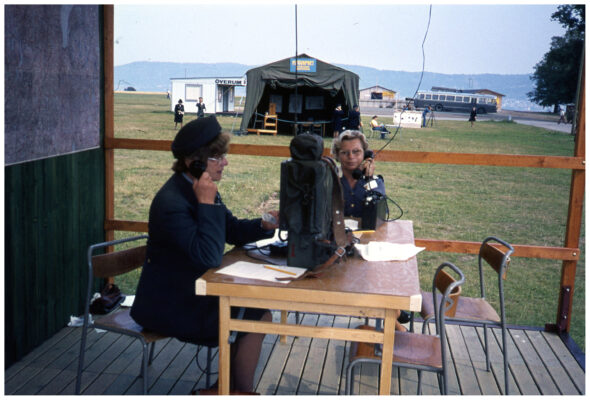 Två kvinnor i uniform sitter vid ett bord med radioutrustning på ett trädäck. I bakgrunden syns ett militärtält. 