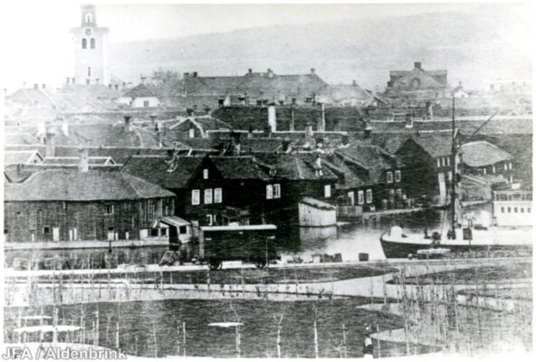 Trähus längs vatten, inklusive en vagn och en segelbåt. 