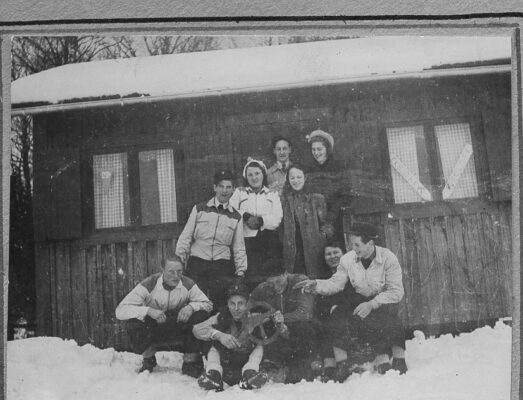 Tio vinterklädda ungdomar framför en stuga. Det är snö på marken.