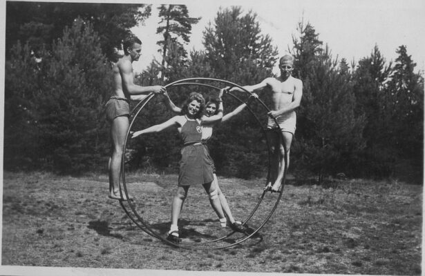 Två unga män stående på sidorna av ett stort metallhjul. I hjulet står två kvinnor.