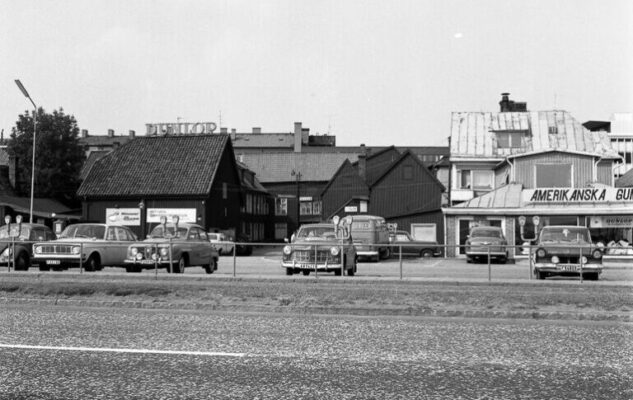 Parkering med ett antal bilar framför några hus.