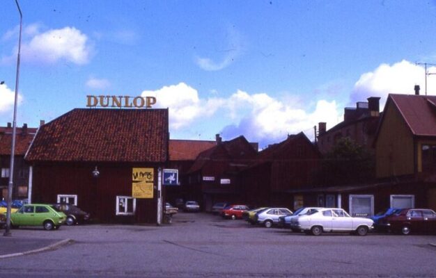 Parkering och bakom flera trähus varav ett med stor skylt "Dunlop" på taket.