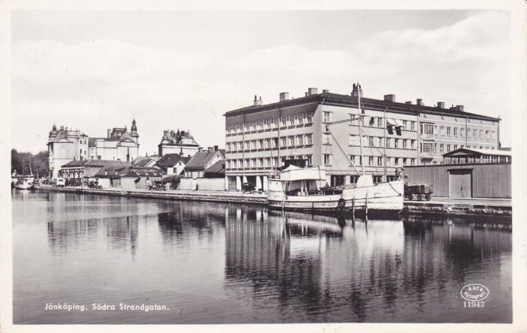 Stort fyrkantigt hus med många fönster på avstånd, sedd från över sjön med en båt framför.