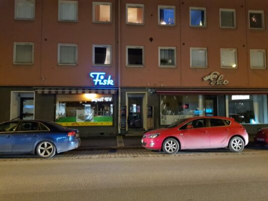 Två butiker sedda från andra sidan gatan, inklusive två parkerade bilar, skylt för "Fisk".
