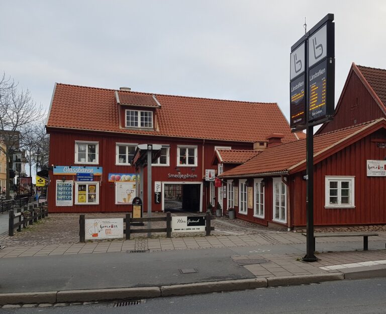 Två sammanbyggda röda trähus, entré till innegården med galler, skylt "Smedjegården", skylta runtom för glass, "Glassbaren", "Gamla Wok", med mera, framför till höger är en busshållplats.
