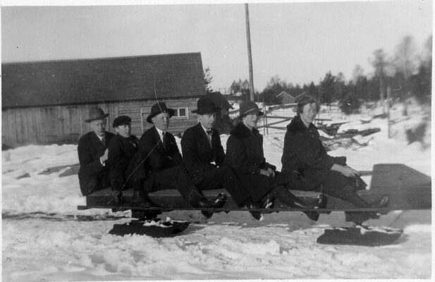 Fyra unga män och två unga kvinnor sittande på en släde i halvprofil. I bakgrunden en mindre lada. 