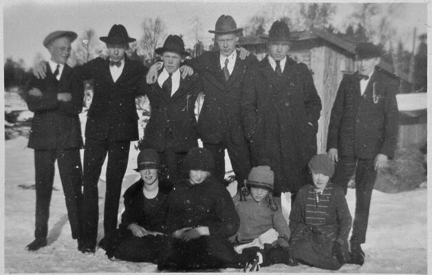 6 unga män stående i kostym. Framför dem 4 kvinnor sittande på snötäckt mark. I bakgrunden ett träskjul.