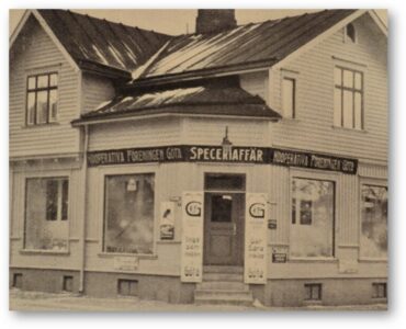 Trähus på två våningar med en diagonal entré i ett av hörnen, skylt "Speceriaffär".