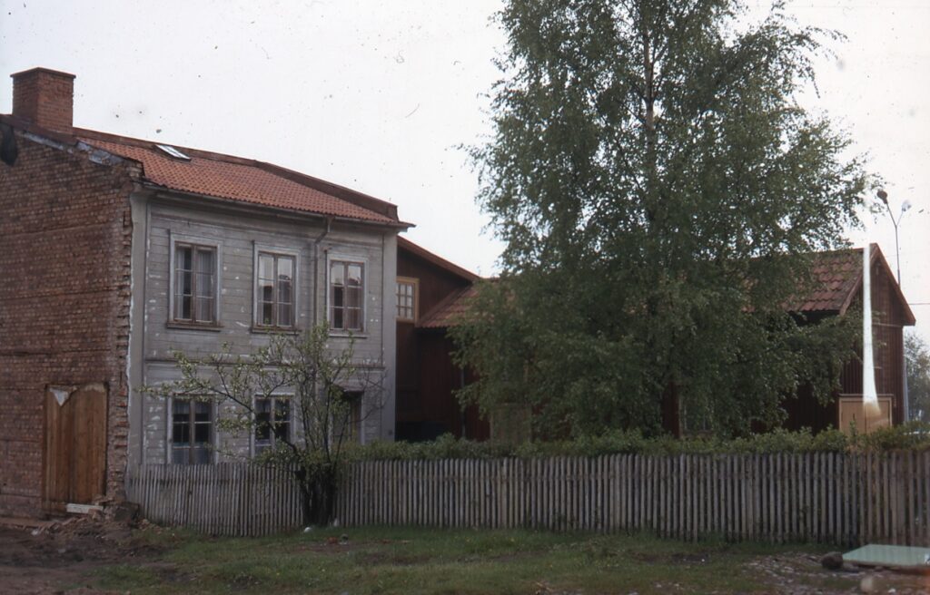 Åldrat trähus med en tegelfasad, övervuxen gård med staket framför.