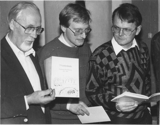 Tre män med glasögon hållande varsitt jubileumsprogram. 