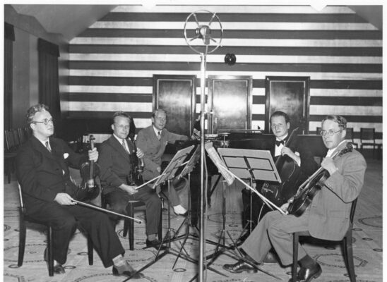 Fem kostymklädda musiker i en studio. Fyra med stråkinstrument, den femte vid en flygel. I förgrunden en mikrofon.