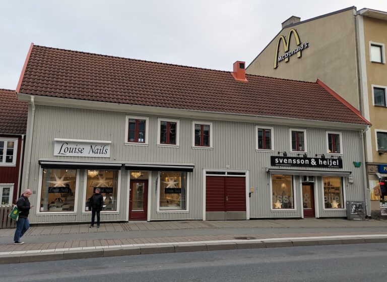 Grått trähus med röda accenter, skylt för "Louise Nails" och till höger "Svensson & Heijel", samt ett hög stenhus med skylt för "McDonald's".