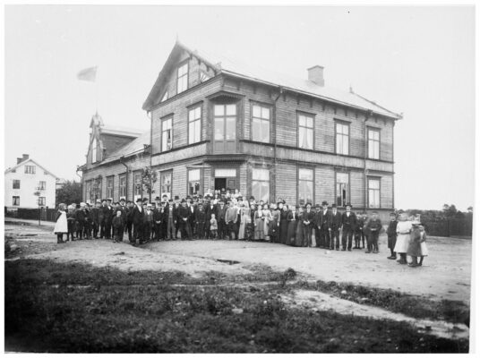 En stor grupp människor – sannolikt logemedlemmarna – samlade framför byggnaden som är ett trevåningshus i trä med en hissad unionsflagga.