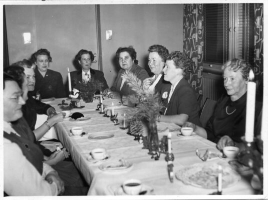 Utklädda och uppklädda damer sitter vid ett bord och har fest.