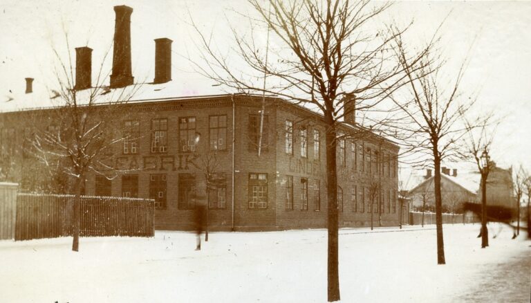 Tegelbyggnad en vinterdag, flera stora skorstenar och texten "Lindells Wågfabrik" på fasaden i stora bokstäver., samt suddiga skepnader av människa och häst på gatan. 