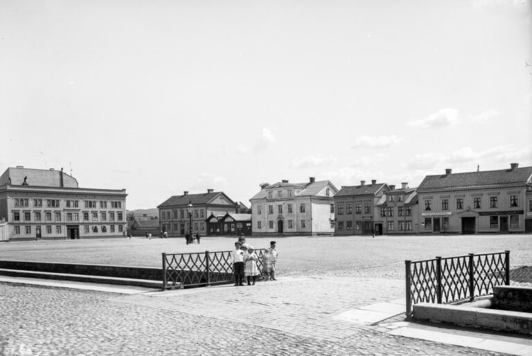 Några barn på bro över kanalen, bakom dem ett stort tomt torg omgivet av byggnader. 
