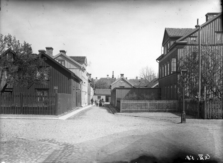 Mynningen till smal sidogata mellan två längor trähus, personer på avstånd.