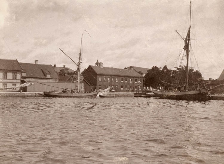 Sjöbild med två båtar, mellan dem syns på avstånd ett avlångt hus på två våningar.