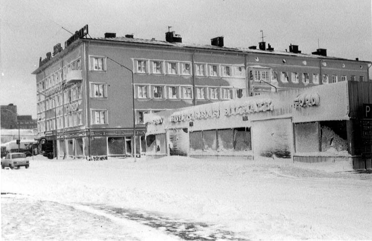 Till höger ett lågt och väldigt insnöat hus, till vänster ett stort fyrkantigt hus med många fönster och några skyltar på taket.