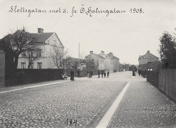 Bred stenbelagd gata, stort trähus till vänster bakom ett staket, med ett antal personer i mitten, till höger ett plank.
