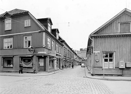 Trähus på hörn vid stengata, bil och kvinna, guldsmedsbutik på hörnet.