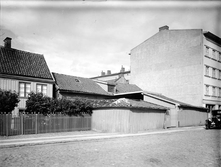 Två trähus med staket och buskar mot en stengata, till höger ett högt stenhus och en bil.