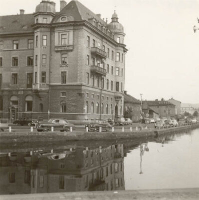 Hörnbild av stort stenhus över sjökanten och en rad bilar som står parkerade invid den, spegelbild i sjön. 
