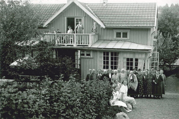 Ett tjugotal människor i en grönskande trädgård framför en trävilla.