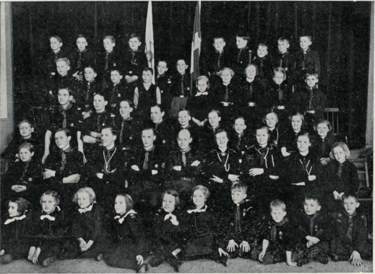 Ett fyrtiotal personer i scoutdräkt, varav flertalet barn, uppställda för fotografering framför ett draperi.