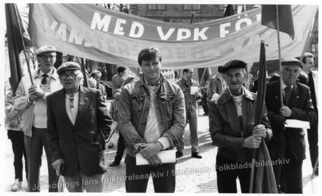 En samling män står framför en fana med text för Vänsterpartiet. 