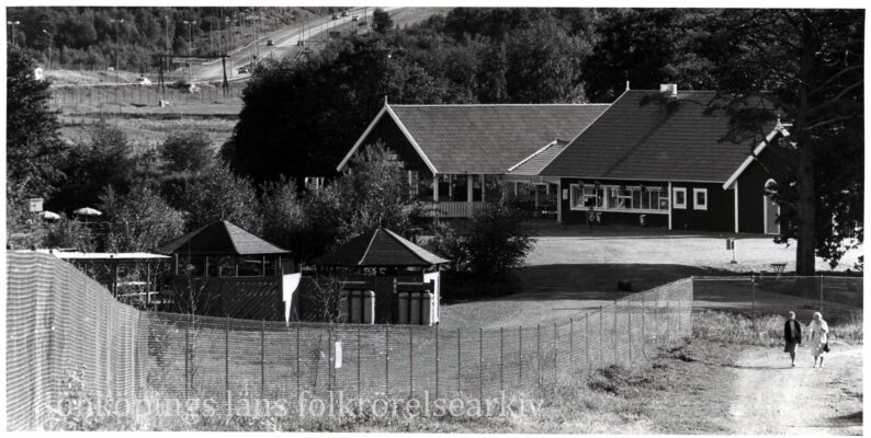Två stugor och paviljonger på avstånd; Till vänster en bilväg och till höger en promenadväg med två gående damer.