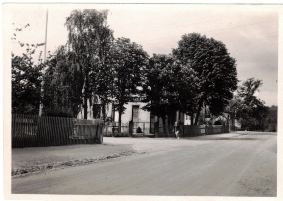 Väg med gårdar som har staket till vänster, ett hus skymtar på en av gårdarna från bakom flera träd.