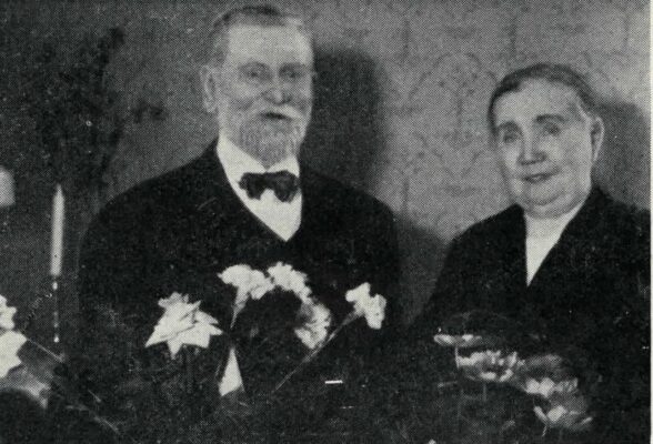 En äldre man i skägg och en äldre kvinna. I förgrunden två blomsterbuketter.