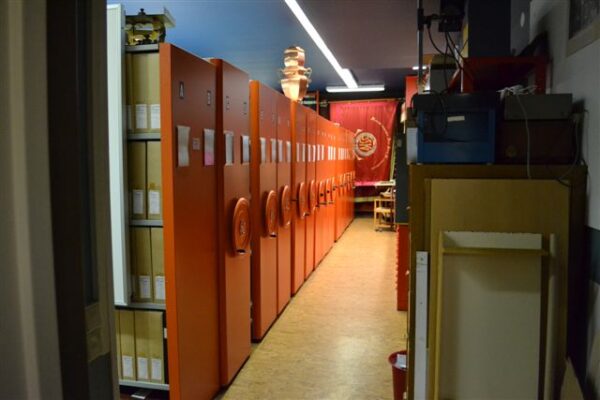 Röda arkivhyllor med arkivboxar på, längst bort i korridoren en röd fana. 