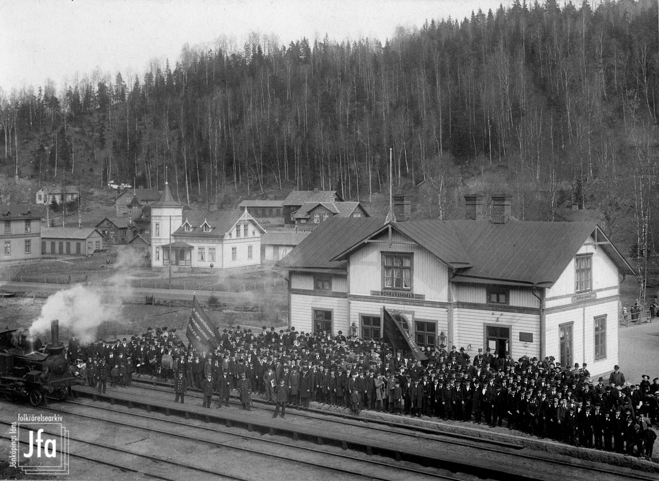 Tågstation i trä med stor samling finklädda herrar med fanor utanför vid och på tågrälsen, till vänster syns ett lok.