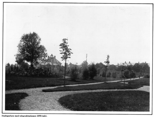 Del av trädgård eller park med prydliga gångstråk. I bakgrunden skymtar ett hus omgärdat av grönska.