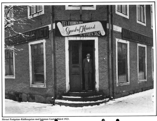 En man i kostym stående vid en gavelentré till ett stenhus. Ovanför dörren en skylt med namnet "Gustaf Håård"