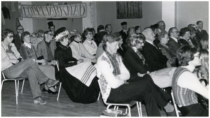 Ett trettiotal personer sitter på stolar i en sal. En del förefaller utklädda i ålderdomliga kläder. I bakgrunden en skylt med texten: "Loppmarknad".