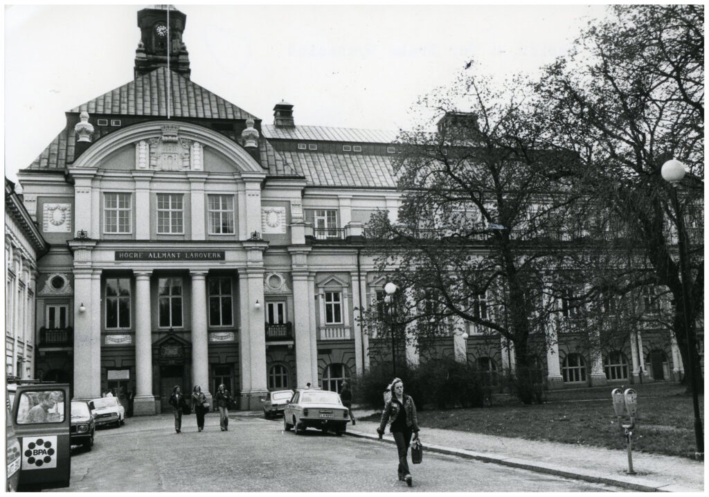 Stor stenbyggnad med kolonner och annan dekoration på fasaden, utanför går en handfull ungdomar och bilar står parkerade.