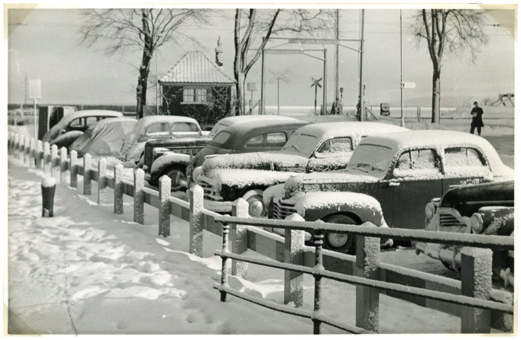 En rad bilar av äldre modell parkerade och täckta av snö en kort bit från en järnvägskorsning.