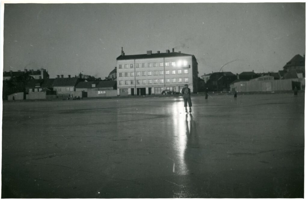 Isäckt sjö med en handfull människor på, stort fyrkantigt hus i bakgrunden.