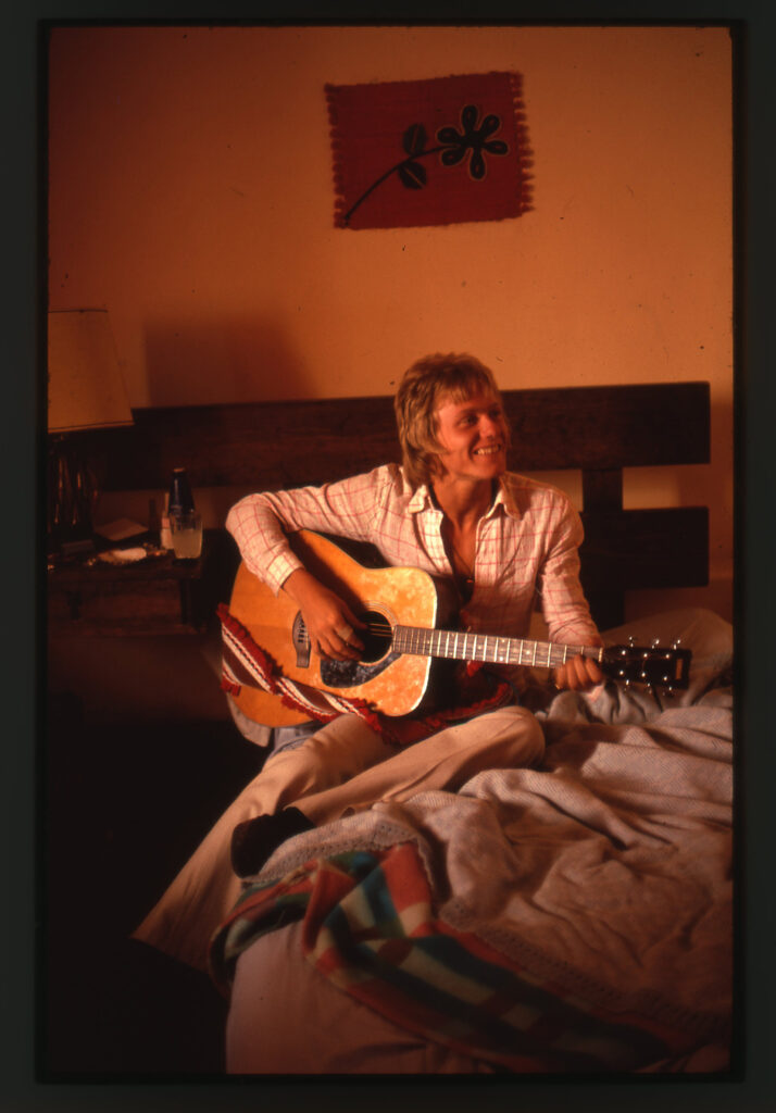 Man med akustisk gitarr på säng.